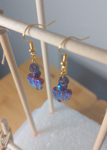 A top-down image of a pair of handmade earrings being displayed on a stand. There is a small, purple, faceted bead on top of a curved triangular bead on top of reflective purple butterflies. The hooks are golden.