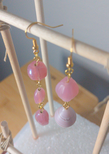 A top-down image of a pair of handmade earrings being displayed on a stand. One earring has a large, pink round bead on top of a thin, oblong, pink bead on top of an irregular shaped, pink bead. The other earring has a large, oblong pink bead on top of an oblong bead with pale pink and white beads.The hooks are golden.