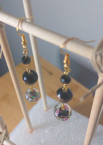A top-down image of a pair of handmade earrings being displayed on a stand. There is a small, black, faceted bead on top of a larger, round black bead with a golden band across the center on top of a black bead with a floral motif. The hooks are golden.