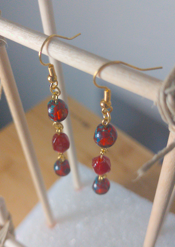 A top-down image of a pair of handmade earrings being displayed on a stand. There is a round red bead on top of a taller diamond shaped red bead on top of another round bead identical to the first. The hooks are golden.