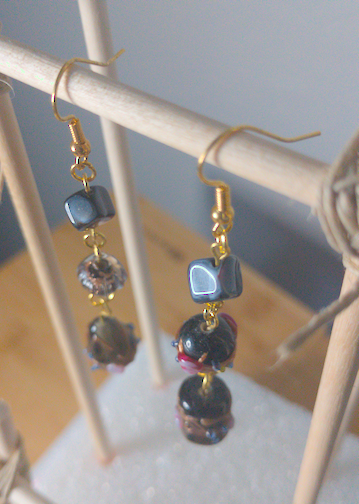 A top-down image of a pair of handmade earrings being displayed on a stand. One earring has a cubic black bead on top of a black bead with a floral motif on top of a tear drop shaped black bead with a floral motif. The other earring has a cubic black bead on top of a round black bead with purple accents on top of a cylindrical black bead with purple and gold accents. The hooks are golden.
