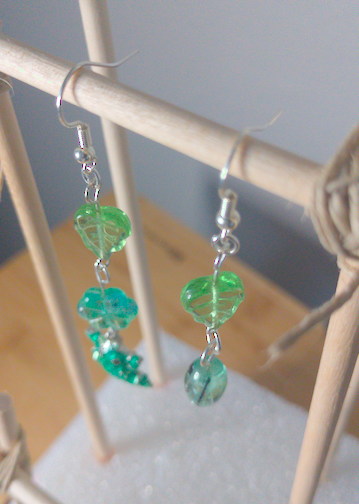 A top-down image of a pair of handmade earrings being displayed on a stand. One earring has a green lead bead on top of a green flower bead. Both beads are on top of a green frog charm. The other earring has a green flower bead on top of a round green bead with black accents. The hooks are a silver color.