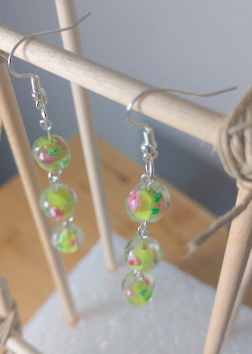 A top-down image of a pair of handmade earrings being displayed on a stand. There are three green bead with a floral motif. The hooks are a silver color.