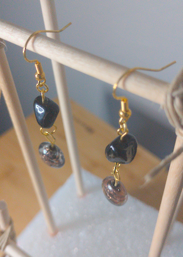 A top-down image of a pair of handmade earrings being displayed on a stand. There is a black, irregular shaped bead on top of a circular black beads with gold swirls. The hooks are golden.
