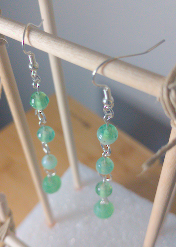 A top-down image of a pair of handmade earrings being displayed on a stand. There are four green beads that slightly increase in the size down the earrings. The hooks are a silver color.
