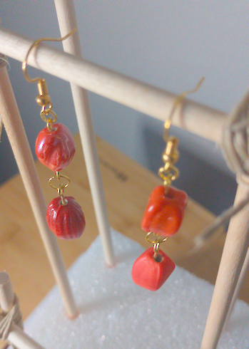 A top-down image of a pair of handmade earrings being displayed on a stand. One earring has a round orange bead with a slight twist on top of an oblong rectangular bead. The other earring has a rectangular orange bead on top of an oblong rectangular bead. The hooks are golden.