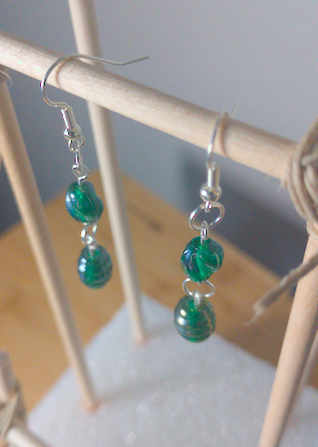 A top-down image of a pair of handmade earrings being displayed on a stand. There is a small, round, green bead with a slight twist on top of a slightly larger green bead. The hooks are a silver color.