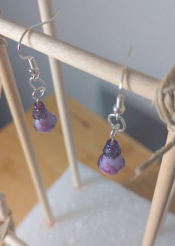 A top-down image of a pair of handmade earrings being displayed on a stand. There is a small, purple seed bead on top of a slightly larger, faceted purple bead on top of a large, purple faceted bead.The hooks are silver.
