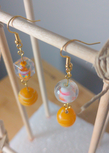 A top-down image of a pair of handmade earrings being displayed on a stand. One earring has a square, clear glass bead with an orange center on top of a large, round, orange bead with white swirls. The other earring has a large, round, clear bead with a white and orange center on top of a large, round, orange bead with white swirls. The hooks are golden.
