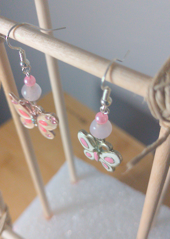 A top-down image of a pair of handmade earrings being displayed on a stand. One earring has a small, round, pink bead on top of a slightly larger, round, white bead on top of a metal butterfly charm. The other earring has a small, round, pink bead on top of a slightly larger, round, white bead on top of a slightly smaller metal butterfly charm. The hooks are silver. 