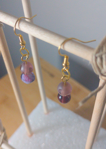 A top-down image of a pair of handmade earrings being displayed on a stand. One earring has a frosted purple bead on top of a larger, faceted, purple bead.The other earring has a frosted purple bead on top of a smooth purple bead. The hooks are golden.