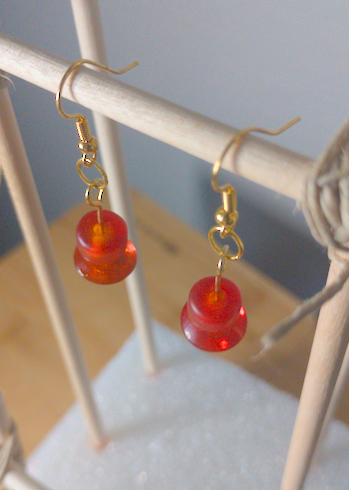 A top-down image of a pair of orange handmade earrings being displayed on a stand. There is a cylindrical bead on top of a disk shaped bead. The hooks are a golden color.