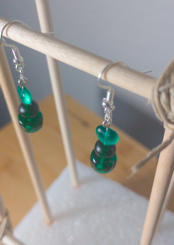A top-down image of a pair of green handmade earrings being displayed on a stand. There is a heart bead on top of a round bead on top of a cylindrical bead. The hooks are a silver color.