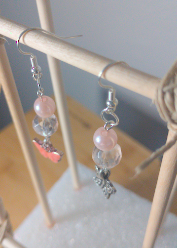 A top-down image of a pair of handmade earrings being displayed on a stand. One earring has a round pink bead on top of a faceted clear bead on top of a metal, orange butterfly charm. The other earring has a round pink bead on top of a faceted clear bead on top of a metal butterfly charm. The hooks are silver. 