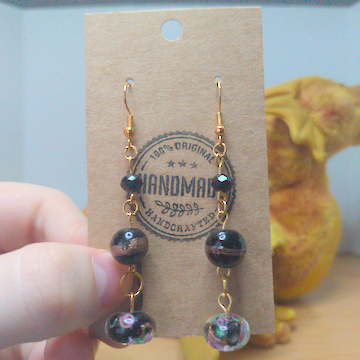 A front facing image of a pair of handmade earrings. There is a small, black, faceted bead on top of a larger, round black bead with a golden band across the center on top of a black bead with a floral motif. The hooks are golden.