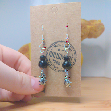 A front facing image of a pair of handmade earrings. There is a small black faceted bead over a larger round black bead. The beads are on top of a metal charm in the shape of a beetle. The hooks are a silver color.