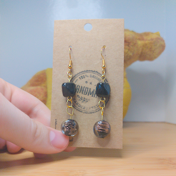 A front facing image of a pair of handmade earrings. There is a black, irregular shaped bead on top of a circular black beads with gold swirls. The hooks are golden.
