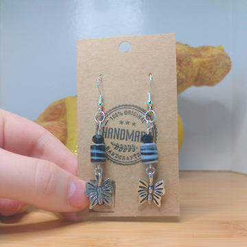 A front facing image of a pair of handmade earrings. There is a small black faceted bead on top of a cylindrical bead with black and white swirls. Both beads are on top of metal charms in the shape of butterflies. The hooks are a silver color.
