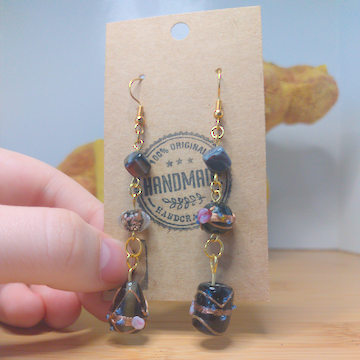 A front facing image of a pair of handmade earrings. One earring has a cubic black bead on top of a black bead with a floral motif on top of a tear drop shaped black bead with a floral motif. The other earring has a cubic black bead on top of a round black bead with purple accents on top of a cylindrical black bead with purple and gold accents. The hooks are golden.