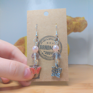 A front facing image of a pair of handmade earrings. One earring has a round pink bead on top of a faceted clear bead on top of a metal, orange butterfly charm. The other earring has a round pink bead on top of a faceted clear bead on top of a metal butterfly charm. The hooks are silver. 