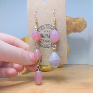 A front facing image of a pair of handmade earrings. One earring has a large, pink round bead on top of a thin, oblong, pink bead on top of an irregular shaped, pink bead. The other earring has a large, oblong pink bead on top of an oblong bead with pale pink and white beads.The hooks are golden.