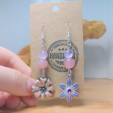 A front facing image of a pair of handmade earrings. One earring has a purple, iridescent bead on top of a pink bead on top of a pink metal flower charm. The other earring has a purple iridescent bead on top of a pink bead on top of a purple metal flower charm. The hooks are silver. 