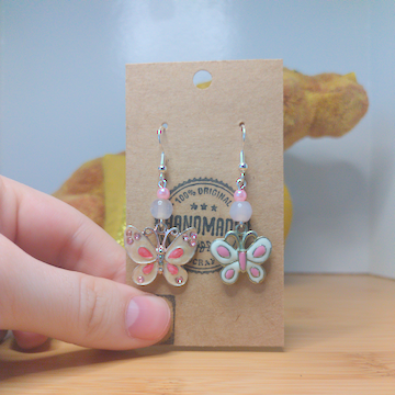 A front facing image of a pair of handmade earrings. One earring has a small, round, pink bead on top of a slightly larger, round, white bead on top of a metal butterfly charm. The other earring has a small, round, pink bead on top of a slightly larger, round, white bead on top of a slightly smaller metal butterfly charm. The hooks are silver. 