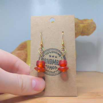 A front facing image of a pair of orange handmade earrings being displayed on a stand. There is a cylindrical bead on top of a disk shaped one. The hooks are a golden color.