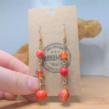 A front facing image of a pair of orange handmade earrings. There is are three round beads of increasing size going down the earring. The hooks are a golden color.