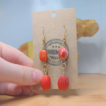 A front facing image of a pair of handmade earrings. One earring has around orange bead with a slight twist on top of an oblong rectangular bead. The other earring has a rectangular orange bead on top of an oblong rectangular bead. The hooks are golden.