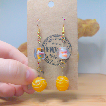 A front facing image of a pair of handmade earrings. One earring has a square, clear glass bead with an orange center on top of a large, round, orange bead with white swirls. The other earring has a large, round, clear bead with a white and orange center on top of a large, round, orange bead with white swirls. The hooks are golden.