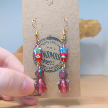 A front facing image of a pair of handmade earrings. There is a small, red, star shaped bead on top of a small, red, diamond shaped bead on top of a small, round, red bead on top of a larger, oblong red bead with small nobs. The hooks are golden.
