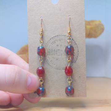 A front facing image of a pair of handmade earrings. There is a round red bead on top of a taller diamond shaped red bead on top of another round bead identical to the first. The hooks are golden.