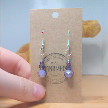 A front facing image of a pair of handmade earrings. There is a small, purple seed bead on top of a slightly larger, faceted purple bead on top of a large, purple faceted bead.The hooks are silver.