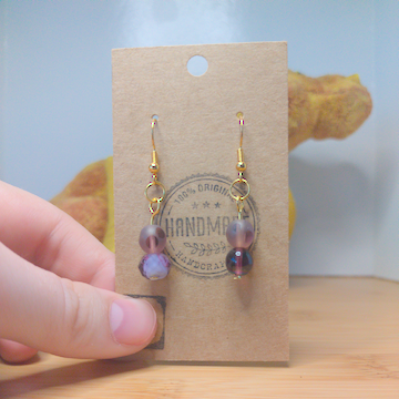 A front facing image of a pair of handmade earrings. One earring has a frosted purple bead on top of a larger, faceted, purple bead.The other earring has a frosted purple bead on top of a smooth purple bead. The hooks are golden.