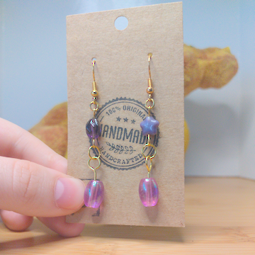A front facing image of a pair of handmade earrings. One earring has a dark purple diamond shaped bead on top of an oblong purple bead. The other earring has a purple star shaped bead on top of an oblong purple bead. The hooks are golden.