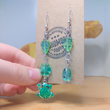 A front facing image of a pair of handmade earrings. One earring has a green leaf bead on top of a green flower bead. The final bead on this earring is a green frog charm. The other earring has a green leaf on top of a round green bead with black accents. The hooks are a silver color.