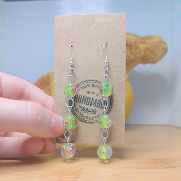 A front facing image of a pair of handmade earrings. There three stacked green beads with a floral motif. The hooks are a silver color.