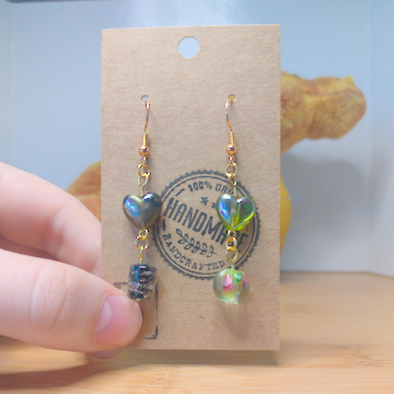A front facing image of a pair of handmade earrings being displayed on a stand. One earring has a dark green heart bead over a black cylindrical bead with gold swirls. The other earring has a lighter green heart earring with a round green bead with a floral motif. The hooks are a golden color.