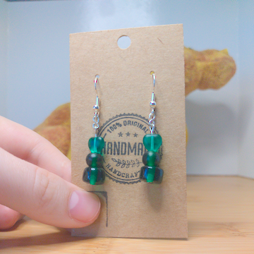 A front facing image of a pair of handmade earrings. There are three green beads stacked. One is a heart, one is round, and one is a cylinder. The hooks are a silver color.