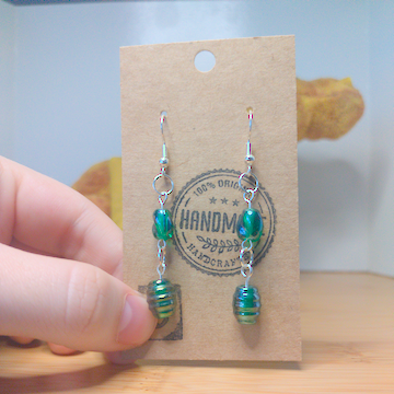 A front facing image of a pair of handmade earrings. There is a small round green bead with a slight twist. It is on top of a slightly larger green bead. The hooks are a silver color.