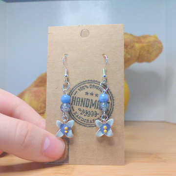 A front facing image of a pair of handmade earrings. There is a light blue bead on top of a cracked glass bead. Both beads are on top of a blue flower charm. The hooks are a silver color.