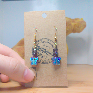 A front facing image of a pair of handmade earrings. There is a small, purple, faceted bead on top of a curved triangular bead on top of reflective purple butterflies. The hooks are golden.